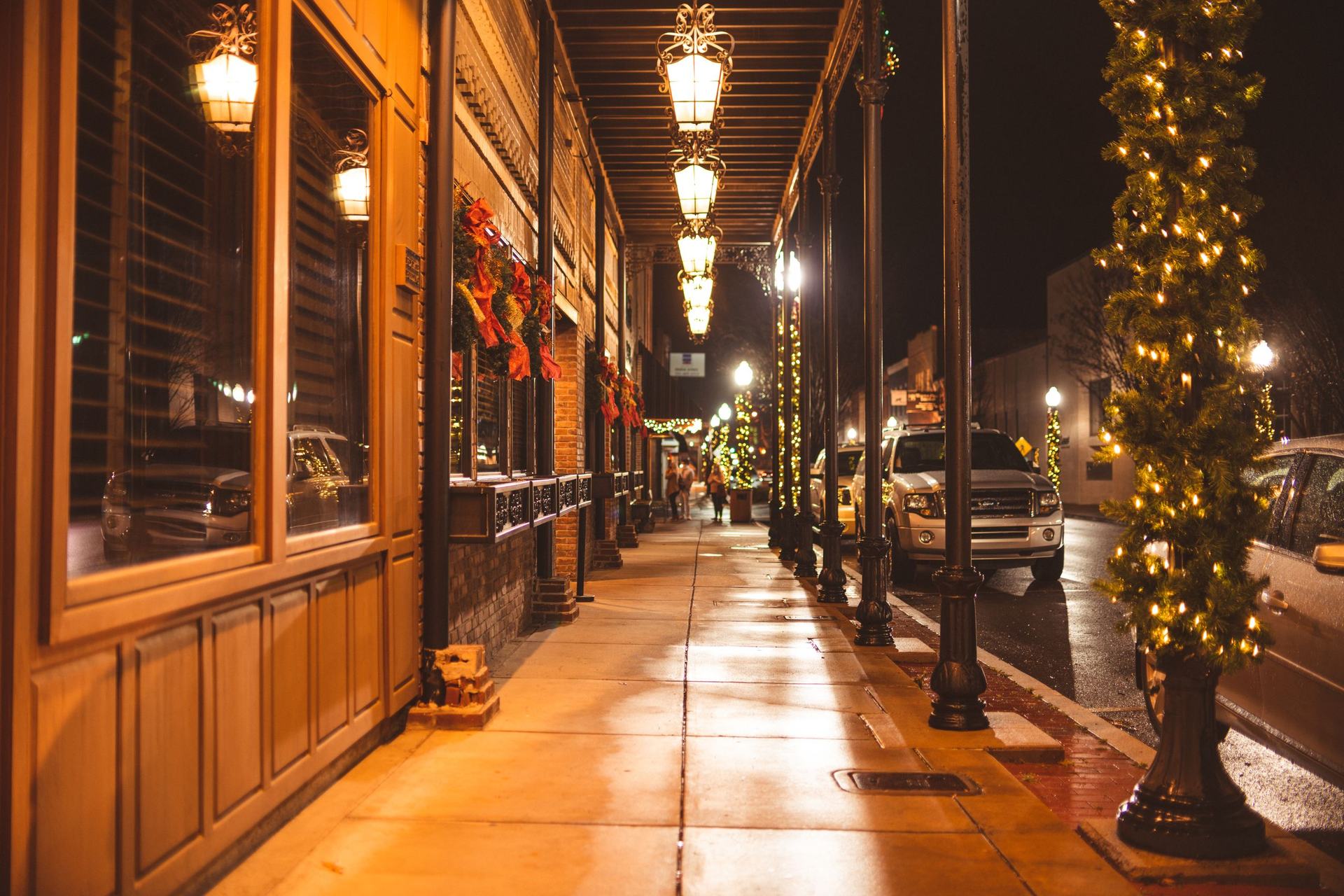 Covered sidewalk along a historic brick storefront at night, hung with lanterns, wreaths and garlands of Christmas lights