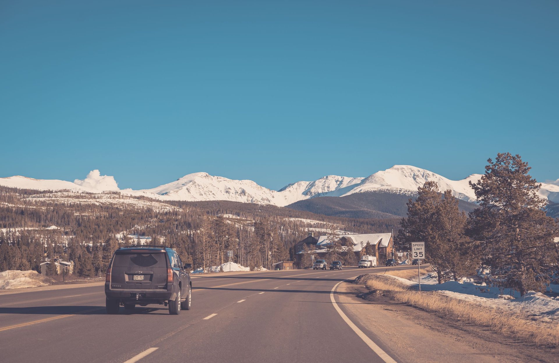 SUV driving a curving two-lane mountain highway toward snow-capped peaks, with snow banks along the roadside