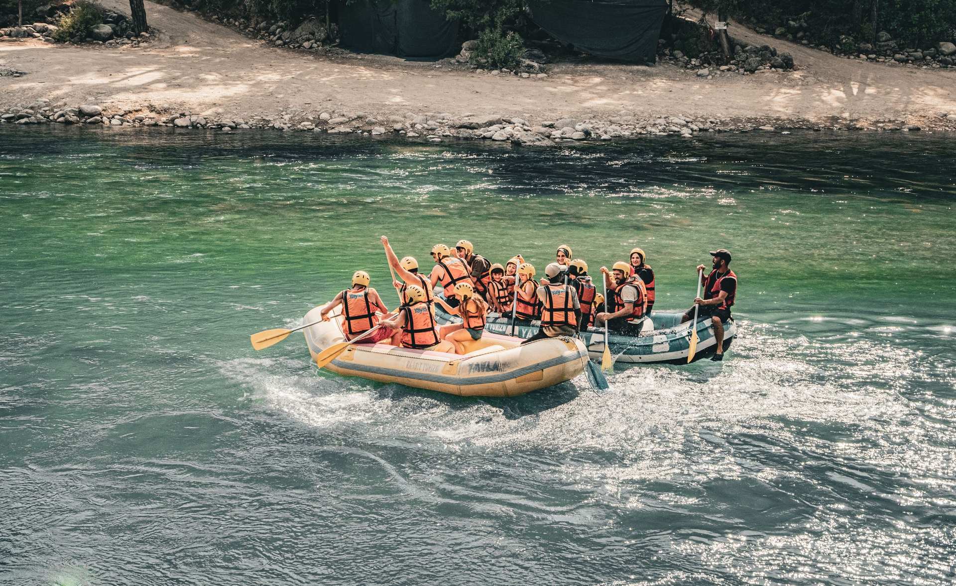 Raft full of paddlers in orange life jackets crossing green river water beside a gravel bank