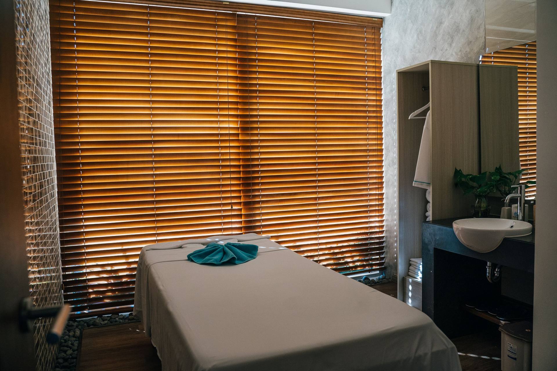 Massage table with a folded teal towel in a quiet treatment room lit through wooden window blinds