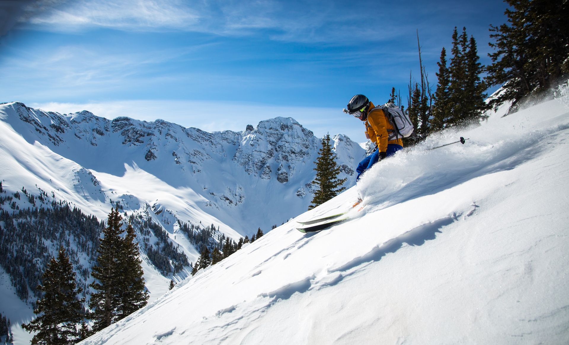 Skier descending a steep, untracked snow slope with jagged snow-covered peaks and spruce trees behind