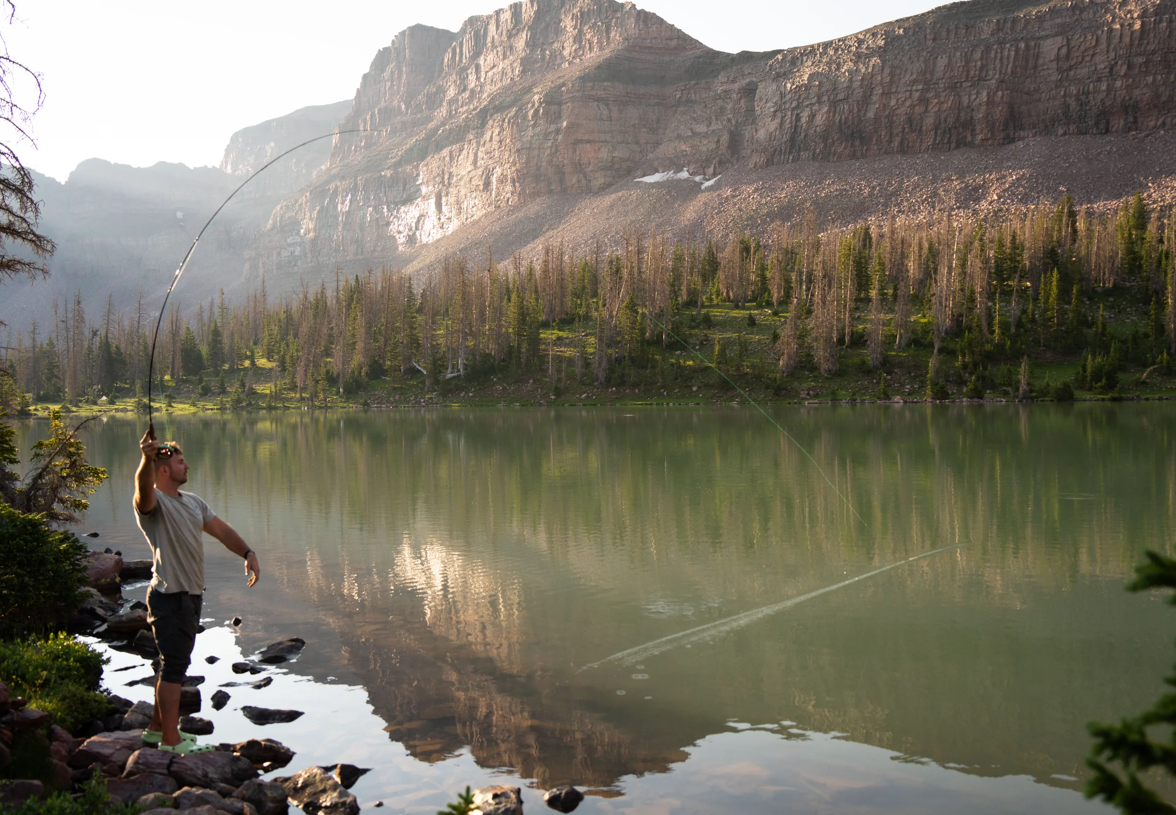 An angler casting a fly rod from the shore of a calm alpine lake below rocky cliffs