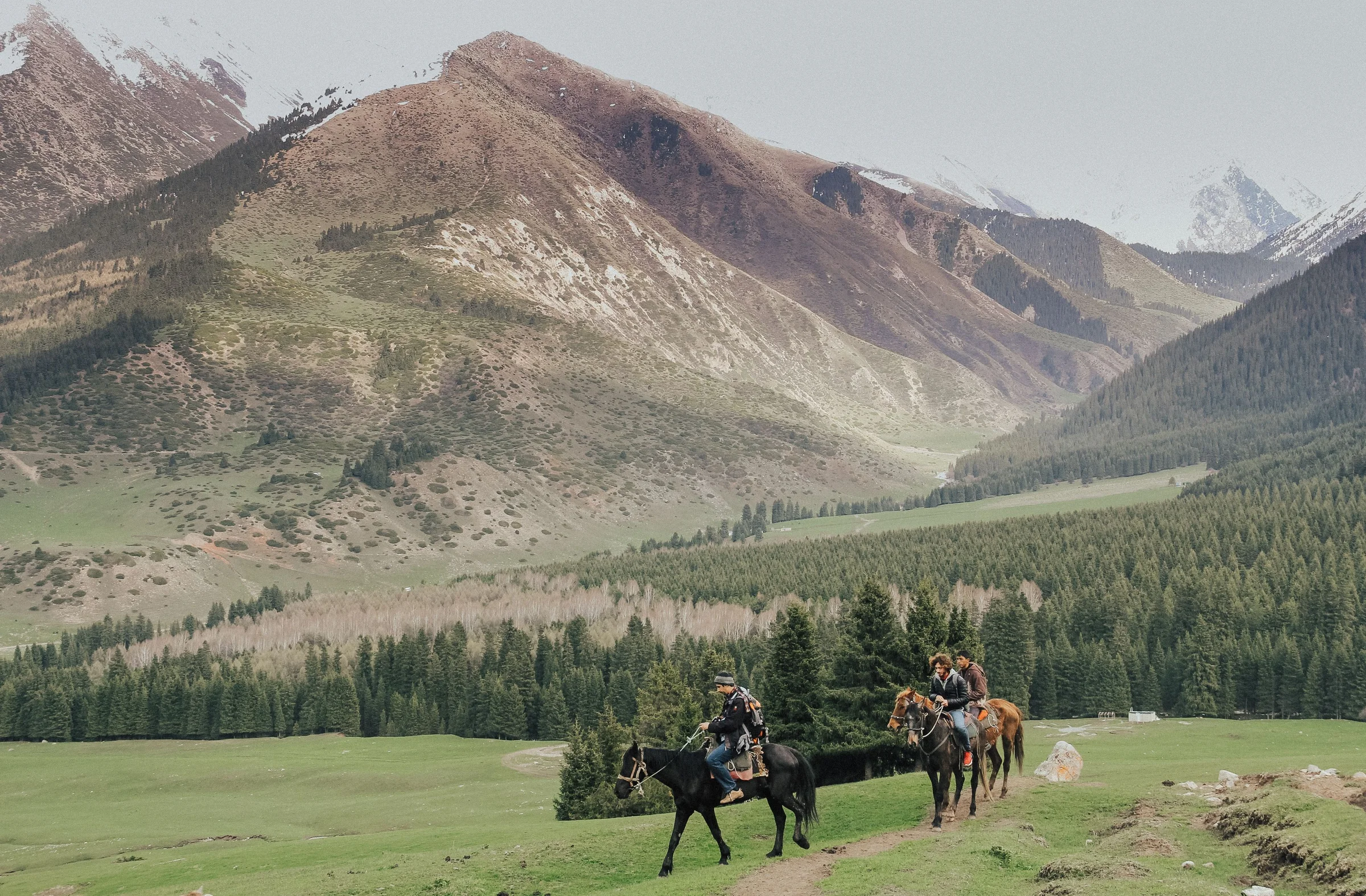 Two riders on horseback crossing a green mountain valley beneath rugged peaks
