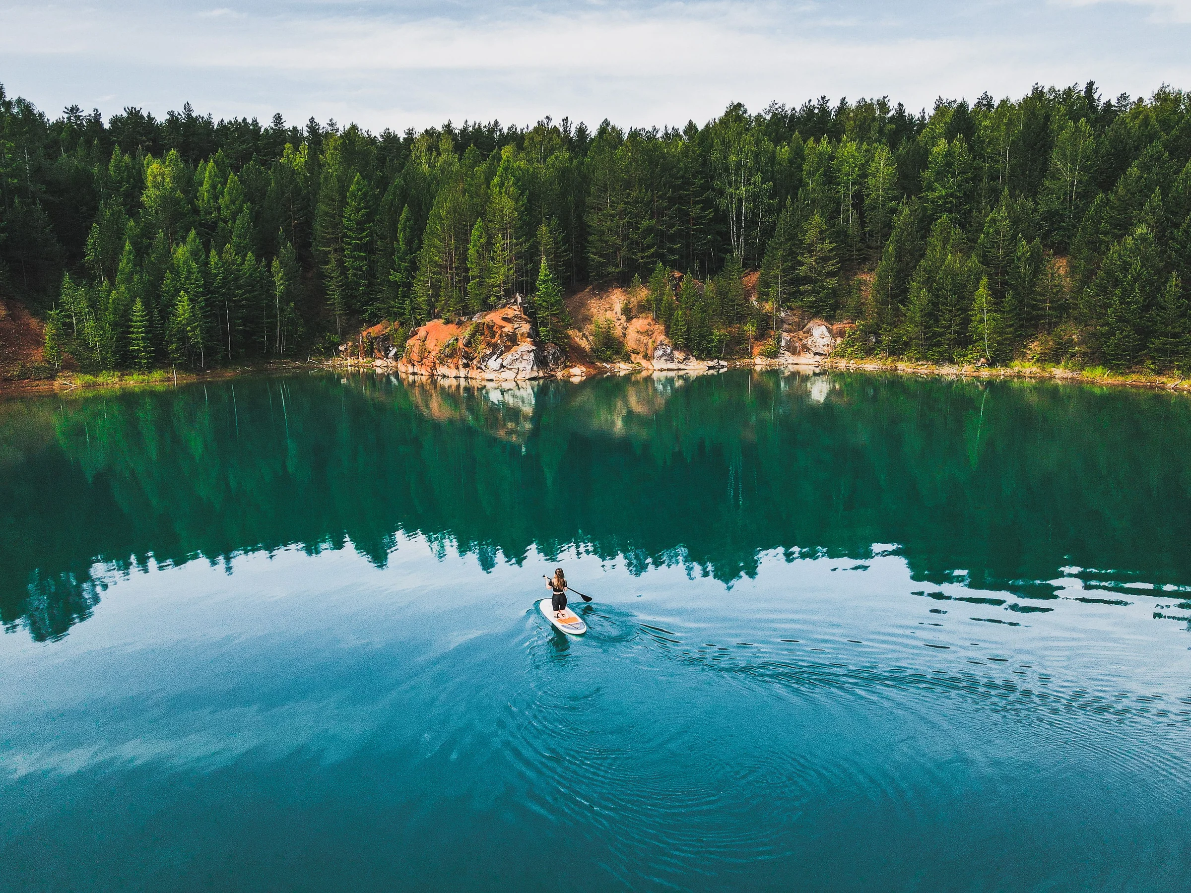 A paddleboarder on a calm lake ringed by forest, seen from above